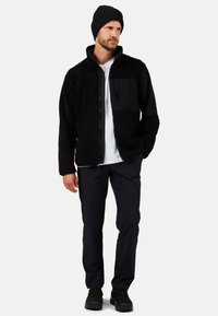 Czarna polarowa kurtka z gładką czarną wstawką na piersi, noszona na białej koszuli, w zestawieniu z ciemnymi spodniami i czarną czapką beanie.