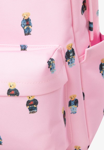 Sac à dos rose avec un tissu à motifs représentant de petits ours en peluche vêtus de différentes tenues. Comprend une poche avant et une texture lisse.