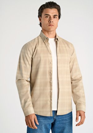 Beige karierter Langarmhemd mit Knopfleiste und leicht strukturiertem Stoff, getragen über einem weißen T-Shirt, kombiniert mit blauen Jeans.