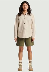 Camisa bege de manga longa com botões e colarinho, combinada com calções verdes-oliva e botins cremosos até ao tornozelo. Tecido texturizado e botões visíveis.