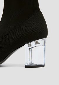 Botte de cheville en tricot noir avec un talon bloc transparent. Le talon présente un design géométrique et une finition lisse et brillante.