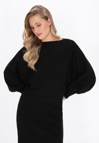 Schwarzer gestrickter Pullover mit übergroßen Ärmeln und einem breiten Halsausschnitt, kombiniert mit einem taillierten schwarzen Rock. Glatte Textur, minimalistisches Design.