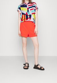 Camicia a maniche corte colorata con bottoni e motivi geometrici in blu, rosso, giallo e bianco; abbinata a pantaloni corti a vita alta rossi brillanti. Sandali neri.