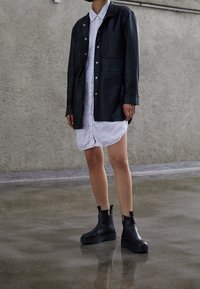 Czarna skórzana kurtka z guzikami, zestawiona z białą lnianą sukienką z guzikami z przodu. Czarne botki za kostkę z złotymi zamkami błyskawicznymi.