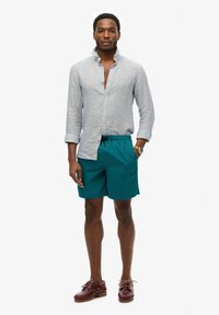 Lichtgrijze linnen shirt, met een open kraag, gedragen over teal zwemshorts met een logo, aangevuld met bordeauxleren bootschoenen.