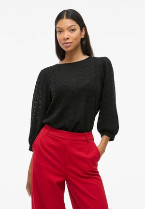 Blouse noire texturée avec un motif en chevron, col rond et manches bouffantes longues, associée à un pantalon rouge taille haute.