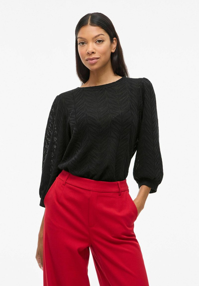 Blouse noire texturée avec un motif en chevron, col rond et manches bouffantes longues, associée à un pantalon rouge taille haute.