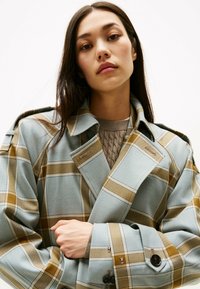 Gabardina cruzada con patrón a cuadros en azul claro y marrón; presenta un gran cuello, ajuste relajado y detalles de botones decorativos en las mangas.