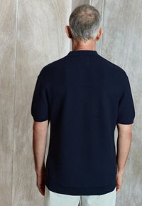 Polo majica kratkih rukava u tamnoplavoj boji od teksturiranog materijala. Ima rebrasti ovratnik i rub, s opuštenim krojem i jednostavnim dizajnom.