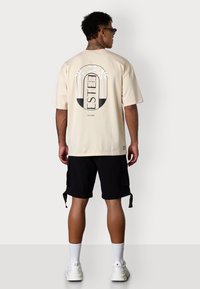 Hellbeige übergroßes T-Shirt mit einem Grafikdesign von Palmen und dem Wort "ESTEEM" in Schwarz. Getragen mit schwarzen Cargo-Shorts.