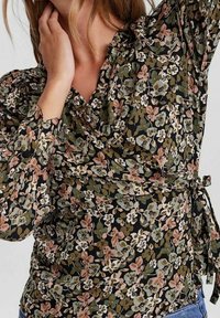 Blouse à motifs floraux avec un fond noir orné de fleurs vertes, roses et blanches. Manches puff longues et détail noué à la taille.