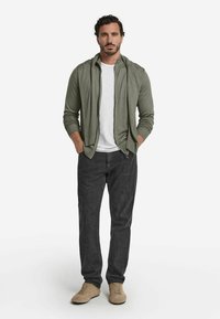 Ljus olivgrön zip-up hoodie över en vit skjorta, parat med mörkgrå jeans och beige skor. Slätt tyg och standard passform.