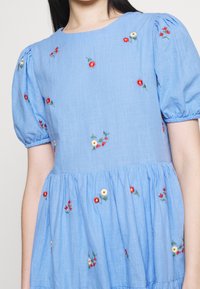 Mujer que lleva un vestido azul claro con mangas cortas abullonadas y bordados de pequeñas flores rojas, blancas y amarillas esparcidos por la tela.