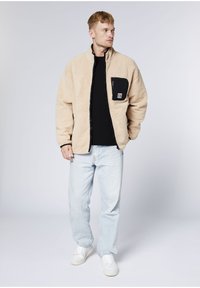 Beige Fleecejacke mit einer schwarzen Reißverschlusstasche und hohem Kragen. Trägt darunter ein schwarzes Hemd mit hellblauen Jeans und weißen Turnschuhen.