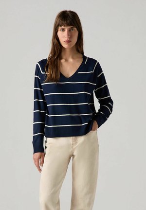 Maglione a righe navy con scollatura a V e maniche lunghe. Presenta righe orizzontali color crema e orlo a coste. Abbinato a pantaloni di colore chiaro.