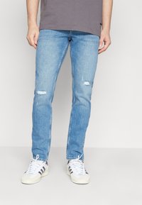 Ljusblå denimjeans med slitna knädetaljer, rak passform och en ren finish, kombinerade med vita sneakers och en grå t-shirt.