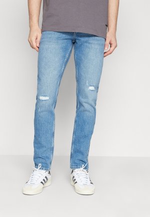 Vaqueros de mezclilla de color azul claro con detalles desgastados en las rodillas, corte recto y un acabado limpio, combinados con zapatillas blancas y una camiseta gris.