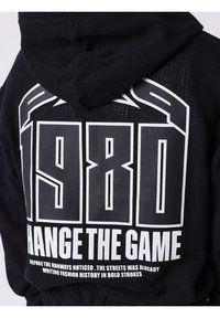 Sweat à capuche noir avec grands textes blancs "1980" et "CHANGE THE GAME" dans le dos, avec un texte plus petit sur l'histoire de la mode en dessous.