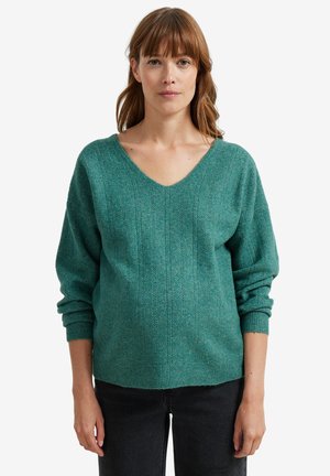 Woman with light brown hair wears teal V-neck long-sleeve sweater and black pants, standing against a plain white background.