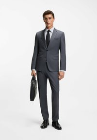 Grauer Anzug mit schmal geschnittenem Design, Ein-Knopf-Verschluss, kombiniert mit einem gemusterten Hemd und schwarzer Krawatte. Schwarze Lederschuhe und eine strukturierte schwarze Tasche.