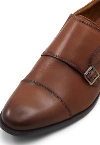 Zapato de vestir de cuero marrón con punta afilada, doble hebilla, acabado suave y costuras contrastantes a lo largo de los bordes.