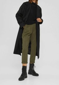 Czarny długi płaszcz na czarnym swetrze, zestawiony z oliwkowozielonymi spodniami i czarnymi sznurowanymi botkami wojskowymi. Teksturowane tkaniny i dopasowany krój.