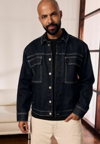Giacca di denim scuro con cuciture bianche, due tasche frontali e bottoni argentati. Indossata sopra una camicia nera, abbinata a pantaloni chiari.