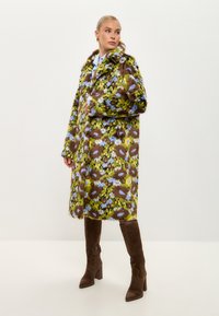 Palton din blană artificială cu model floral în nuanțe de verde, maron și albastru, având guler cu rever și mâneci lungi, purtat cu cizme maron până la genunchi.