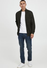 Mørk strikket zipjakke, hvit t-skjorte under, blå jeans og hvite sneakers. Stoffet har en strukturert finish med ribbede detaljer.