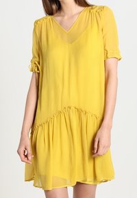 Žluté šaty po kolena vyrobené z lehkého, průhledného materiálu. Mají V-krk, krátké rukávy s mašlemi a nařasený lem.