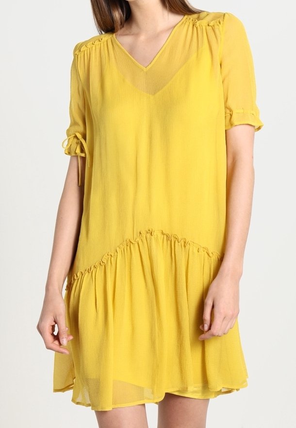 Žluté šaty po kolena vyrobené z lehkého, průhledného materiálu. Mají V-krk, krátké rukávy s mašlemi a nařasený lem.