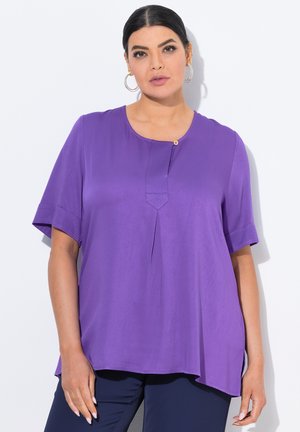 Frau trägt eine locker sitzende lila Bluse mit kurzen Ärmeln und einem einzelnen oberen Knopf, kombiniert mit dunkler Hose, steht vor einem weißen Hintergrund.
