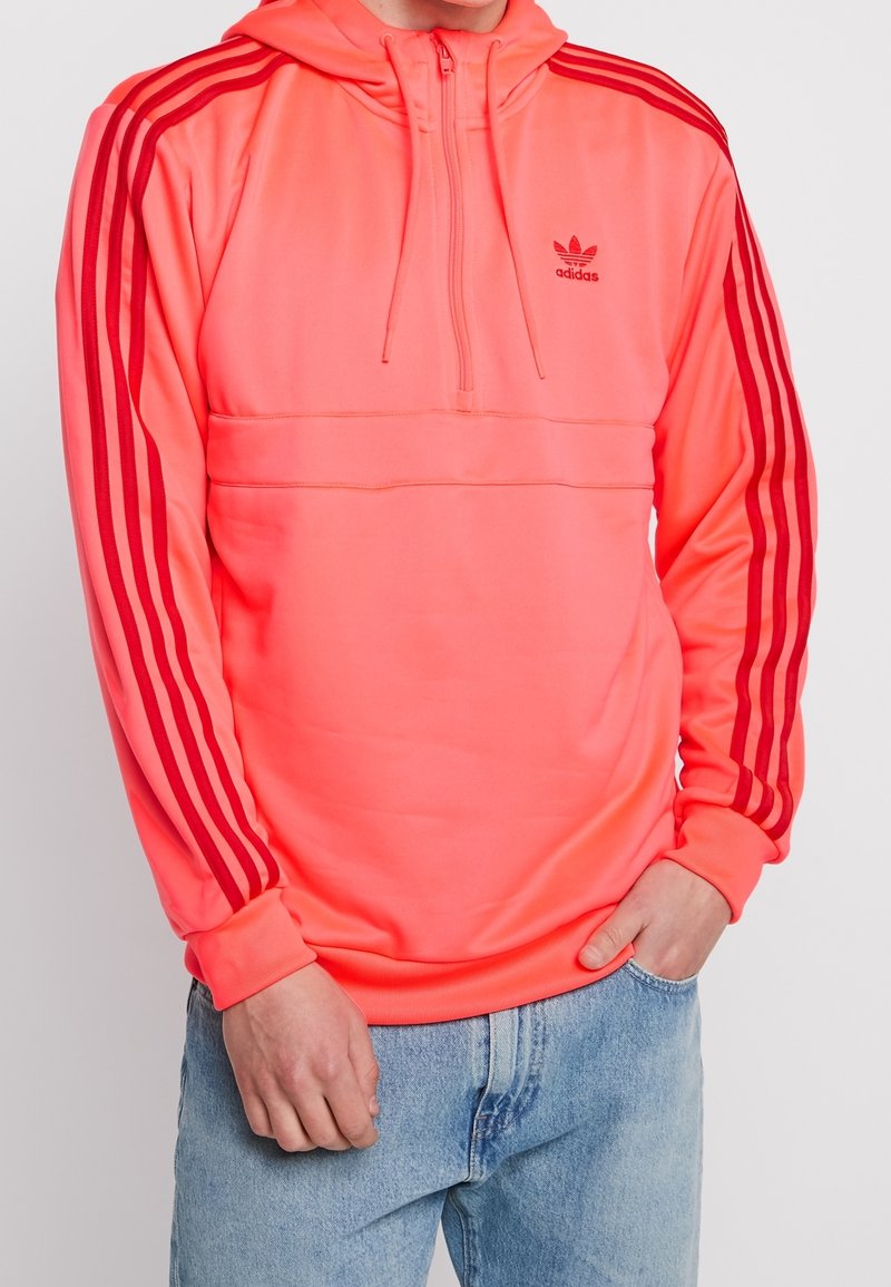 Haut à capuche zippé rose vif avec des accents rouges à trois rayures sur les manches, présentant une poche kangourou et une texture de tissu lisse et extensible.