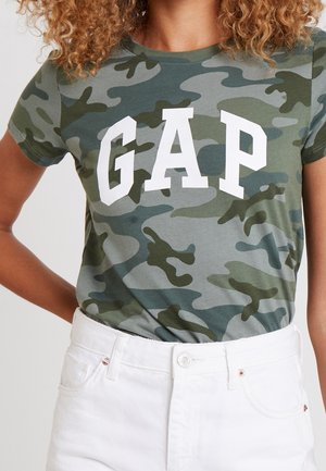 Person trägt ein grünes und graues Camouflage GAP T-Shirt, in hoch taillierte weiße Hose gesteckt, steht mit einer Hand in der Hüfte.