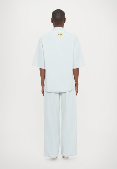 Camisa de ganga azul clara com mangas curtas, colarinho dobrado e um rótulo quadrado na parte de trás. Calças largas e soltas feitas do mesmo material.