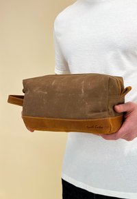 Brown canvas and leather toiletry bag with a zip closure, tan accents, and a side handle. Features a logo embossed on the bottom.