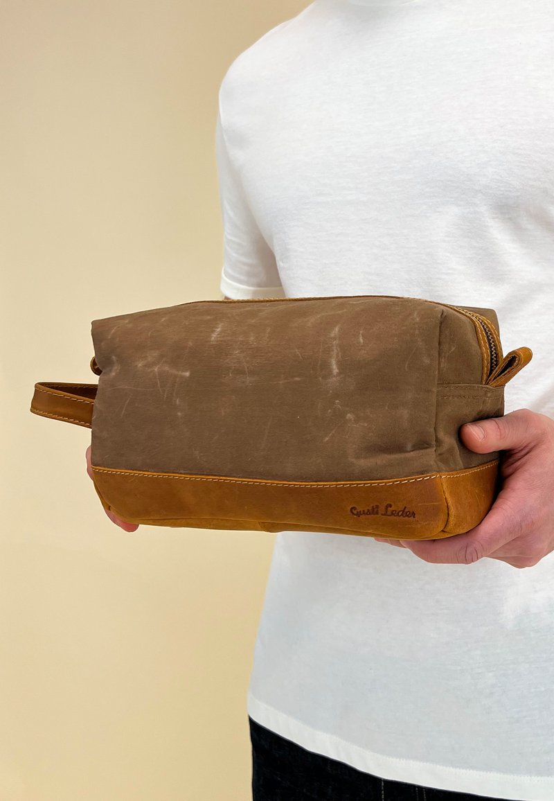 Brown canvas and leather toiletry bag with a zip closure, tan accents, and a side handle. Features a logo embossed on the bottom.