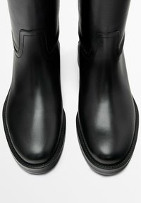 Černé kožené kotníkové boty s hladkou texturou, zaoblenou špičkou, výrazným prošíváním a zakřivenými ozdobami na svršku. Ploché podrážky.