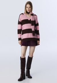 Roze en bruine gestreepte zachte trui met een V-hals en sjaalaccent, gecombineerd met een donkerpaarse rok en bruine kniehoge blokhaklaarzen.