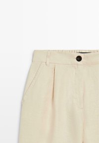 Pantalon beige taille haute avec plis, passants de ceinture, poches latérales et fermeture par un bouton foncé sur un fond plat.