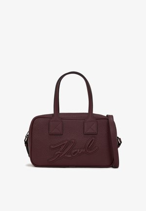 Burgundijas ādas rokassoma ar teksturētu virsmu, kurai ir divi rokturi un regulējams plecu siksna. Uzpriekš ir iespiests uzņēmuma logo.