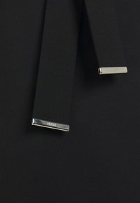 Mustaa kangasta, jossa on kaksi tyylikästä hihnaa, joissa on hopeanvärisiä yksityiskohtia. Toisessa hihnassa on plakaatti, jossa on kaiverrettu "HUGO", mikä korostaa modernia muotoilua.