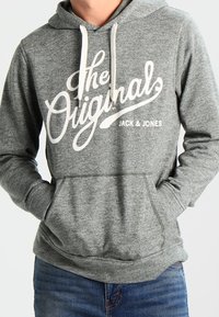 Felpa grigia in cotone con logo bianco stampato "The Originals", con coulisse e tasca frontale, abbinata a jeans blu. Texture morbida, vestibilità regolare.