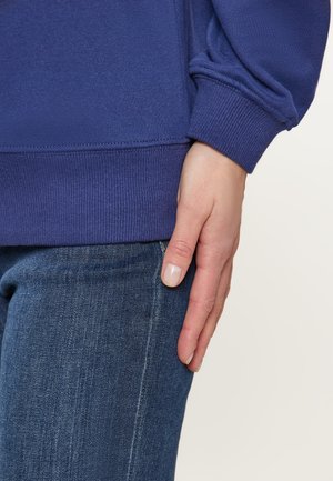 Felpa viola con orlo e polsini a costine, abbinata a jeans in denim blu. La mano poggia sulla coscia, mettendo in mostra una pelle liscia e smalto per unghie neutro.