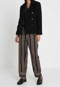 Žena oblečená v černém dvouřadém saku, černých, hnědých a bílých pruhovaných kalhotách a černých mokasínech, stojící na hladkém bílém pozadí.