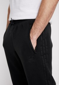 Černé fleecové tepláky se bočními kapsami, s šňůrkou v pase a texturovanými pruhy po stranách. Mají vyšitý logo na stehně.