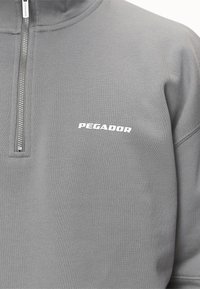 Felpa grigia con zip realizzata in tessuto testurizzato. Presenta un colletto alto e il logo "PEGADOR" in bianco sulla parte sinistra del petto.