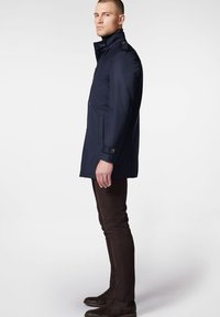 Granatowy wodoodporny płaszcz z wysokim kołnierzem, dwoma bocznymi kieszeniami i zapięciem na guziki. Wykonany z fakturowanej tkaniny, z mosiężnymi akcentami.
