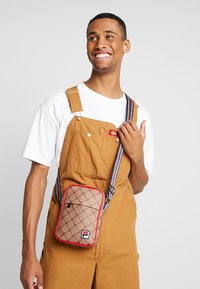 Sac en bandoulière à motifs marron avec des accents rouges et une bandoulière rayée, présentant un détail de logo à l'avant.