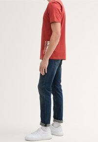 Červené bavlněné tričko s krátkým rukávem a bílým grafickým potiskem na boku, spojené s tmavými džínami a bílými teniskami.